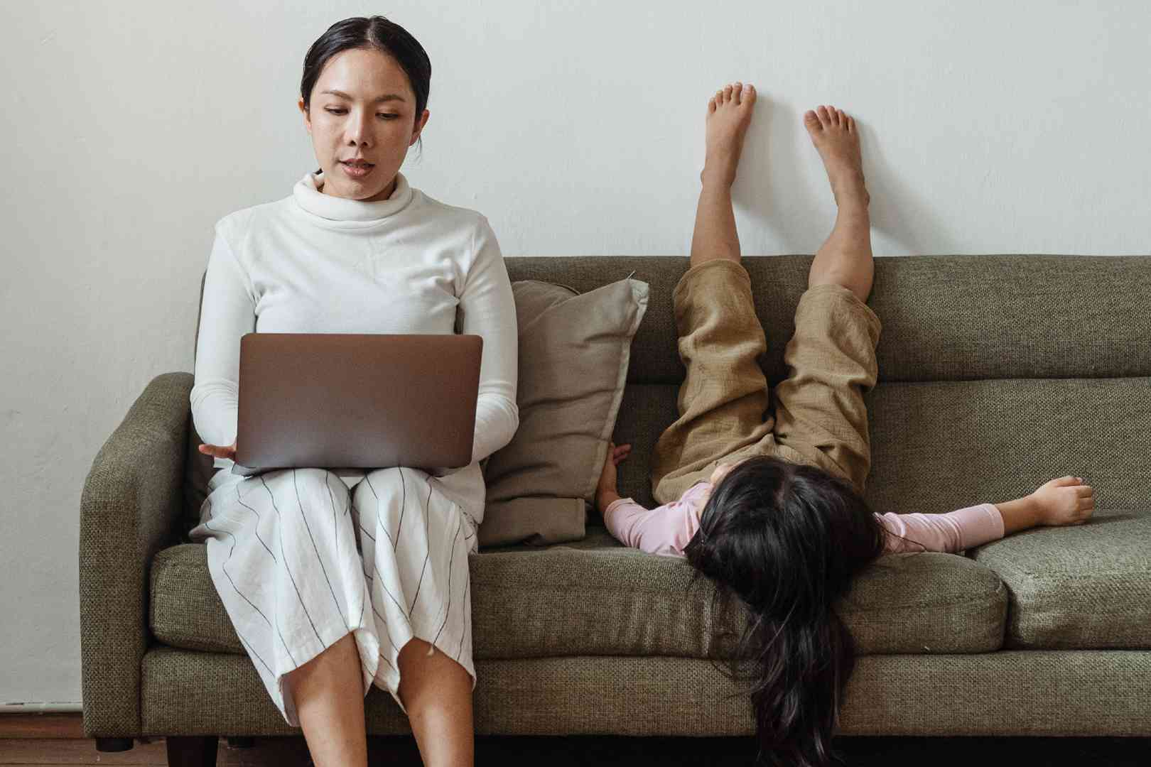 A woman sitting on the couch with her laptop and a child laying down.