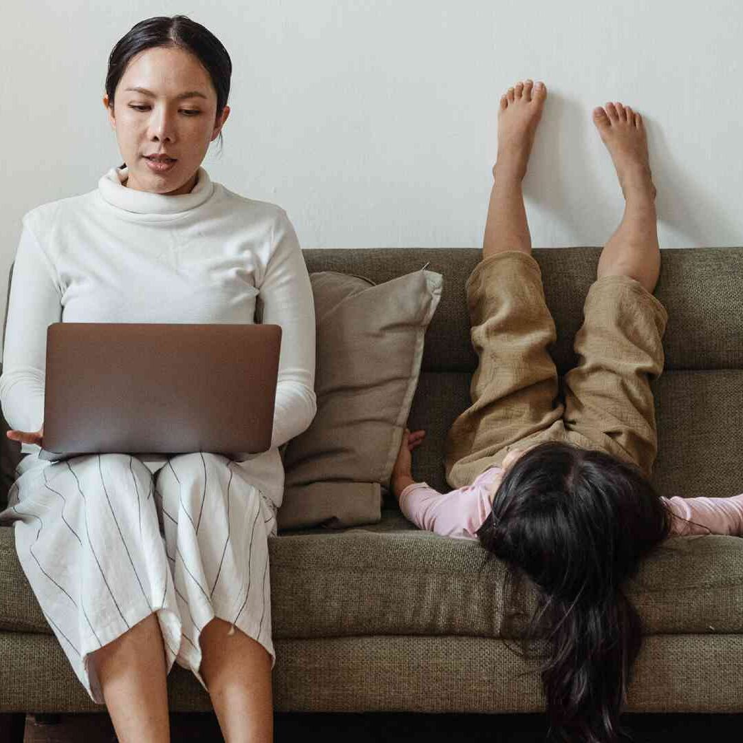 A woman sitting on the couch with her laptop and a child laying down.
